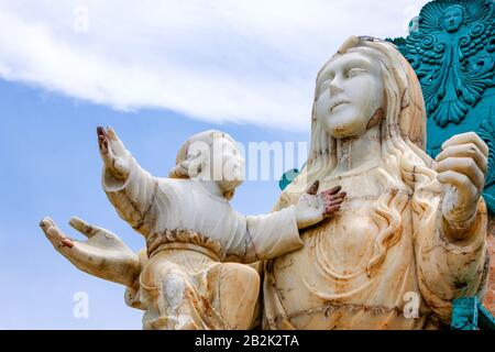 Eine Nahaufnahme einer verwitterten weißen Marmorstatue, die eine Frau mit langen, fließenden Haaren zeigt, die ein Kind hält. Das Kind, das links der Frau steht, hebt eine Hand nach oben, während die andere aufliegt Stockfoto