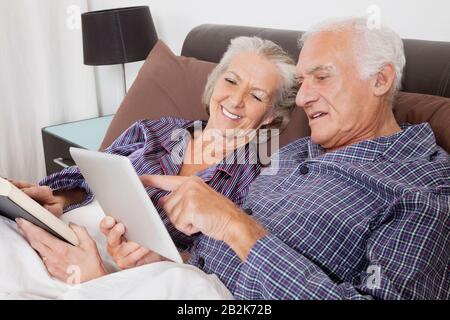 Glückliches älteres Paar, das ein digitales Tablet verwendet, während es auf dem Bett im Zimmer liegt Stockfoto