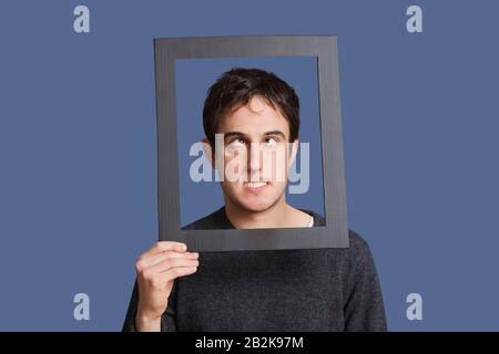 Junger Mann, der lustige Gesichter durch den Rahmen über blauem Hintergrund macht Stockfoto
