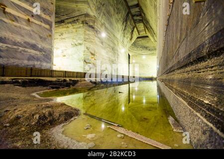 Eine weitläufige unterirdische Salzbergwerkskammer mit hohen, geschichteten Felswänden, die unterschiedliche Schichten in Grau-, weiß- und Blautönen zeigen. Ein flacher Pool aus grüngelbem Wasser reflektiert die Decke Stockfoto