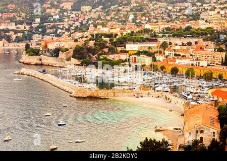 Ein erhöhter Blick auf eine Küstenstadt im mediterranen Stil mit einem geschäftigen Yachthafen voller Segelboote und Yachten. Ein Steinbruch erstreckt sich in das ruhige Meer und schützt den Hafen. Die Stadt steigt auf A Stockfoto