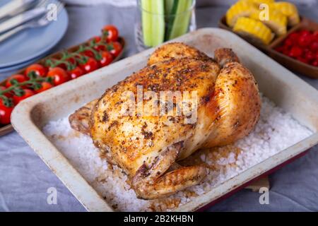 Hähnchen komplett auf Salz gebacken. Mit Mais auf dem Cob, Kirschtomaten, Gurken und Jalapeno-Paprika. Selektiver Fokus, Nahaufnahme. Stockfoto