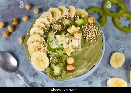 Smoothie-Schüssel aus grünem Obst mit sternförmigen Bananen, Kiwi, Haselnuss und gebucktem Quinoa-Korn auf grauem Grund Stockfoto