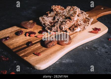 Hausgemachtes Obstbrot auf Holzhackbrett mit verstreuten trockenen Beeren auf dem Hintergrund auf schwarzem Tisch. Stockfoto