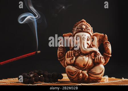 hindu-gott Ganesh auf schwarzem Grund. Rudraksha-Statue und Rosar auf einem Holztisch mit rotem Räucherstäbchen und Räucherstrauch. Kopierbereich Stockfoto