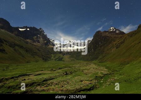 Ein weites Bergtal bei Nacht unter Sternenhimmel mit üppigen grünen Wiesen im Vordergrund und zerklüfteten, felsigen Hängen, die sich auf beiden Seiten erheben. In der Ferne befinden sich schneebedeckte Berggipfel Stockfoto