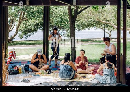 Maroochydore, Australien - 15. Februar 2020. Weibchen weben Körbe auf Handwerkerwerkstatt, sitzen auf Teppich im Park. Vielfalt und Einbeziehung in Kreativität. Frauengemeinschaft. Australien multikulturell. Stockfoto