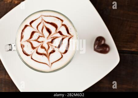 Ein Blick von oben auf eine Kaffeetasse aus Glas, gefüllt mit einem Latte mit komplizierten Blumenkunstwerken, die mit Espresso auf aufgeschäumter Milch zubereitet werden. Der Becher sitzt auf einer weißen quadratischen Platte, begleitet von Stockfoto