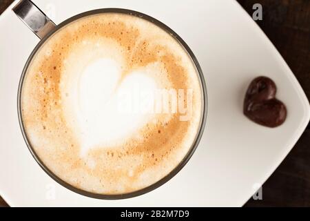 Ein Blick von oben auf einen Latte in einem Glasbecher mit Metallgriff und einem herzförmigen Design im Milchschaum. Der Becher sitzt auf einem weißen quadratischen Teller, begleitet von einer kleinen dunklen Schokolade Stockfoto