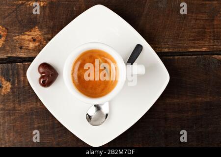 Blick von oben auf eine weiße Espresso-Tasse aus Keramik, gefüllt mit dunklem Kaffee und einer reichhaltigen Crema, auf einer passenden quadratischen Untertasse. Neben dem Becher liegt ein kleines herzförmiges Schokoladenbonbon und ein Metall Stockfoto