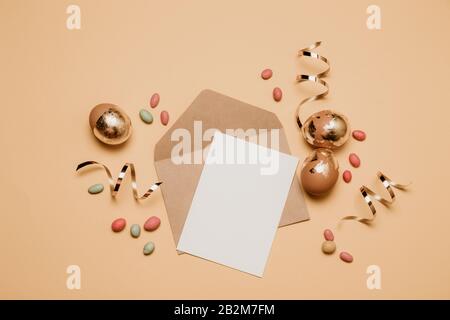 Leerer Umschlag aus Kunsthandwerk mit einem Blatt Papier, goldenem Glitterband auf beigefarbenem Hintergrund. Goldene ostereier mit Gelee Bohnen Süßigkeiten, Draufsicht. Stockfoto