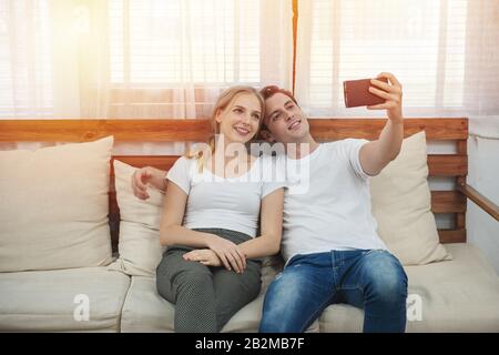 Fröhliches junges kaukasisches Paar sitzt auf der Couch und nimmt selfie zu Hause im Wohnzimmer, romantischer Moment von Mann und Frau in der Freizeit. Stockfoto
