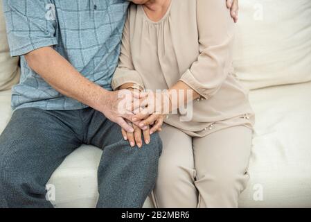 Zugeschnittenes Foto der Hände des süßen älteren asiatischen Ehepaares, das die Hände auf dem Sofa hält.Alte Leute umarmen und halten die Hände. Paarkonzept. Liebevolle Konz Stockfoto