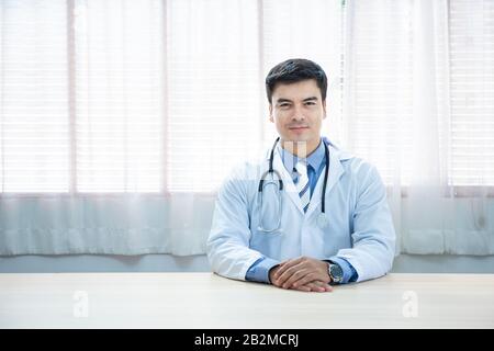 Der junge kaukasische Arzt sitzt am Schreibtisch an seinem Arbeitsplatz und lächelt mit der Kamera. Perfekter medizinischer Service in der Klinik. Stockfoto