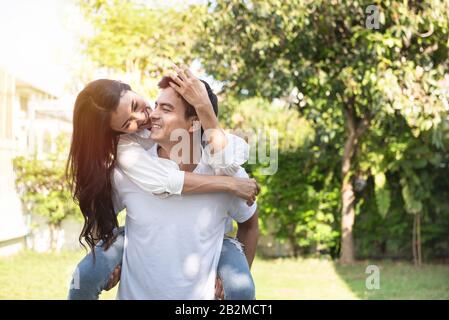 Portrait des jungen Mannes, der seine schöne asiatische Frau auf dem Rücken im Park trägt, der Freund, der im Sommer seiner schönen Freundin Huckepack mitgibt Stockfoto