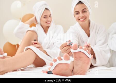 Vorderansicht zweier lächelnder weiblicher Freunde mit Handtüchern am Kopf und in Bademänteln, die auf dem Bett sitzen und Pediküre machen. Selektive Fokussierung der Beine mit nassem Nagel Stockfoto