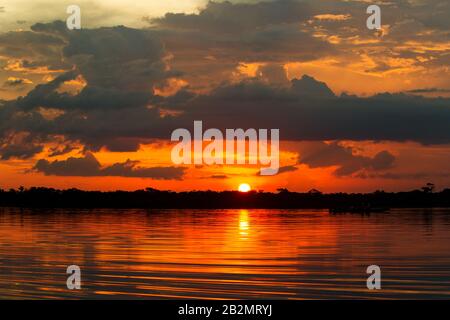 Ein lebhafter Sonnenuntergang über einem ruhigen Gewässer, wobei die Sonne teilweise über einem dunklen Baumbestand am Horizont sichtbar ist. Der Himmel ist voller dramatischer Wolken, die in Orange-, Rot- und Farbtönen beleuchtet sind Stockfoto