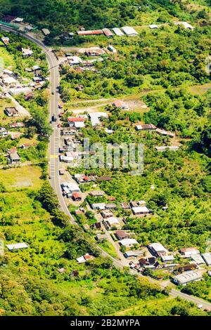 Ein Luftbild zeigt eine gewundene zweispurige Straße, die durch eine ländliche Landschaft mit üppigen grünen Hügeln und verstreuten Häusern führt. Die Straße biegt sich sanft durch das Gelände, begrenzt von dichtem Stockfoto