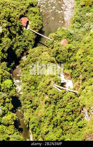 Eine Luftperspektive zeigt eine schmale Hängebrücke, die einen felsigen Fluss in einem dichten, grünen Wald überspannt. Zwei kleine Gebäude mit roten Dächern sind auf beiden Seiten des Flusses zu sehen, die durch die miteinander verbunden sind Stockfoto
