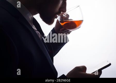 Nahaufnahme, auf einem weißen Hintergrund, die Silhouette eines bärtigen gutaussehenden Kerls, der Tee aus einer durchsichtigen Tasse trinkt, verwendet ein Smartphone. Aktion Stockfoto