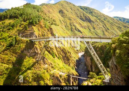 Eine Fußgängerbrücke aus weißem Stahl mit Metallträgern überspannt einen tiefen, schmalen Canyon mit steilen, grünen Felswänden. Unten windet sich ein felsiges Flussbett mit fließendem Wasser durch das Tal Stockfoto