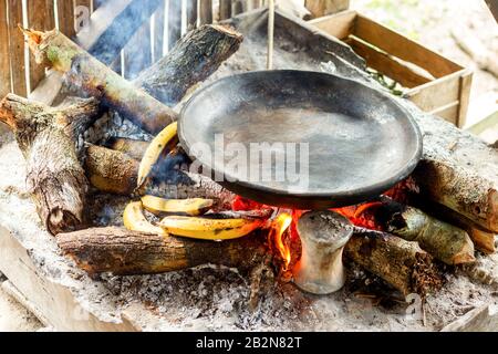 Eine schwarze, runde Eisenpfanne wird über einem Holzfeuer aufgestellt, der von einem Stein- oder Metallständer gestützt wird. Das Feuer brennt hell zwischen geteilten Stämmen und Asche, wobei mehrere gelbe Bananen um den Fuß des angeordnet sind Stockfoto