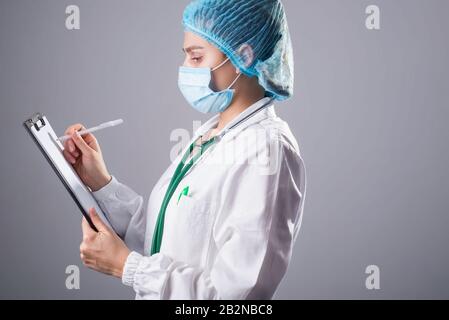 Ein Studio-Porträt eines Arztes in Schutzuniform schreibt medizinische Informationen in einen Ordner. Auf grauem Hintergrund. Ein weiblicher Arzt in einer Schutzmaske Stockfoto