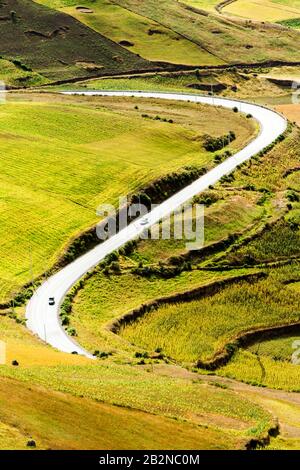 Ein Blick aus der Vogelperspektive auf eine kurvige Asphaltstraße, die durch sanfte grüne Hügel und landwirtschaftliche Felder führt. Die Landschaft zeigt hellgrüne und gelbe Fruchtfelder mit sichtbaren Terrassenmustern Stockfoto
