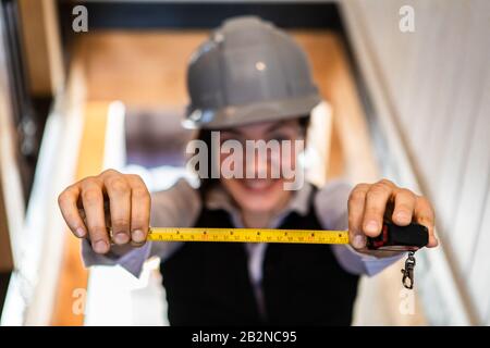 Selektive Fokussierung auf die Hände von Frauen mit einem Metallbandmaß, sie ist eine Architektin, die während der Qualitätskontrolle einen grauen Hardhat im Hintergrund trägt Stockfoto