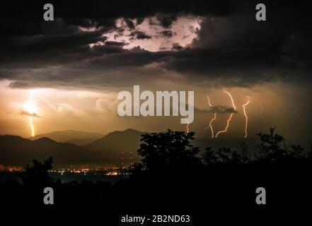 Mehrere helle Blitze beleuchten nachts einen dunklen, stürmischen Himmel über einer Bergkette. Die Blitze zeigen übereinander liegende Wolken und werfen einen goldenen Glanz über den Horizont. Im Stockfoto