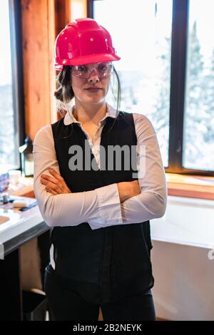 Junge Bauinspektorin, die mit gekreuzten Armen stehen, tragen bei der Luftqualitätsprüfung einen pinkfarbenen Hut auf dem Innenbereich des Hauses. Vertikal Stockfoto