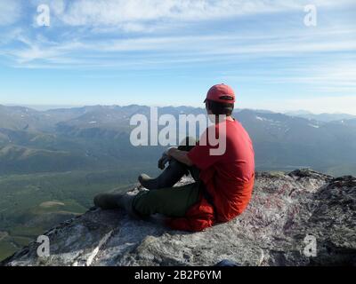 SAMSUNGA-Mann mit rotem T-Shirt und Kappe sitzt in einem Nationalpark im Ural-Gebirge in Russland auf einem Stein auf einem Berg und blickt in die Ferne am Horizont und in die Berge gegen einen blauen Himmel mit Wolken. KAMERABILDER Stockfoto