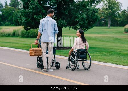Fröhliche behinderte Frau, die einen Ehemann mit Strohkorb betrachtet, während sie im Park mit Babywagen zusammen spazieren geht Stockfoto