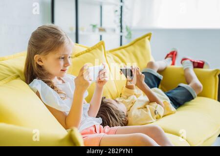 Liebensliches Kind sitzt mit Smartphone in der Nähe eines Bruders auf dem Sofa und benutzt das Smartphone zu Hause Stockfoto