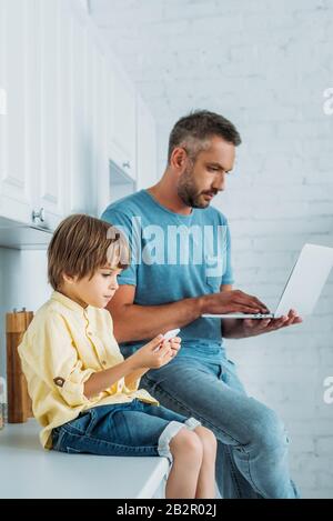 Süßes Kind, das mit einem Smartphome auf der Küchenecke in der Nähe des Vaters sitzt und einen Laptop verwendet Stockfoto