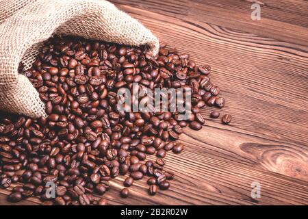 Gebratene Kaffeebohnen, die aus einer Jutekaffeetasche auf einem Holztisch aufwachen. Nahaufnahme. Stockfoto