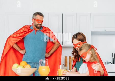 Familie in Superheldenkostümen mit Frühstück in der Küche, während Vater mit Händen auf Hüften steht Stockfoto