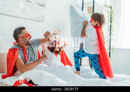 Fröhliche Eltern in Superhelden Kostüme kämpfen mit Sohn, der Kissen hält Stockfoto