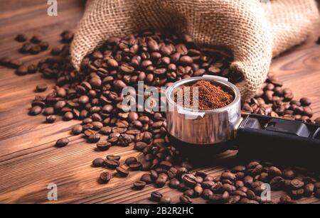Gebratene Kaffeebohnen, die aus einer Jutekaffeetasche aufwachen. Ein porta-filter mit gemahlenem Kaffee auf braunem Holzhintergrund. Stockfoto