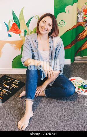 Kaukasische Künstlerin erholt sich, nachdem sie in der Wohnung oder in der Schule Wandbilder mit Acrylfarben in Innenräumen gemalt hat. Kreatives Hobby und freel Stockfoto