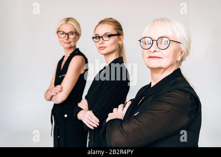 Selektive Fokussierung der modischen, blonden Geschäftsfrauen der drei Generationen in schwarzen Outfits und in grau isolierten Brillen Stockfoto