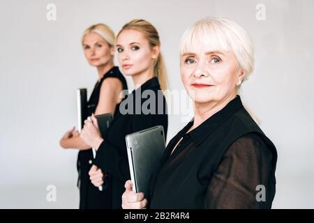 Selektive Fokussierung von blonden Geschäftsfrauen der drei Generation in schwarzen Outfits, die Laptops isoliert in Grau halten Stockfoto