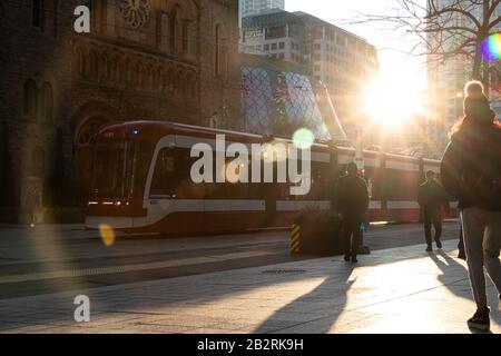 Die Sonne scheint durch die Gebäude in der Innenstadt von Toronto, während eine TTC-Straßenbahn an einem belebten Bürgersteig vorbeifährt. Stockfoto