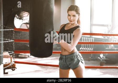 Boxertraining für Frauen in einem Boxring. Boxer übt ihre Bewegungen in einem Boxstudio aus. Das Mädchen umarmt eine Stanztasche und schaut auf die Kamera Stockfoto