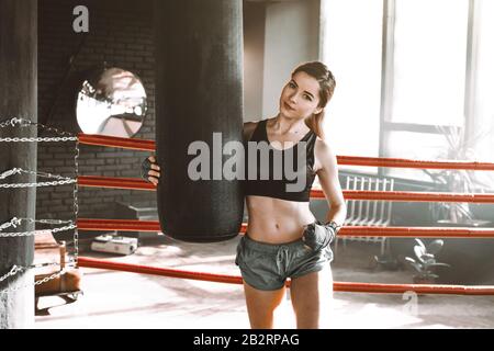Boxertraining für Frauen in einem Boxring. Boxer übt ihre Bewegungen in einem Boxstudio aus. Das Mädchen umarmt eine Stanztasche und schaut auf die Kamera Stockfoto