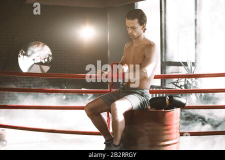 Der junge, gutaussehende Boxer zieht vor dem Kampf oder Training auf dem roten Fass im Boxring. Boxkämpfer Bereiten Verbände Vor Stockfoto