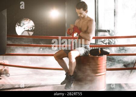 Der junge, gutaussehende Boxer zieht vor dem Kampf oder Training auf dem roten Fass im Boxring. Boxkämpfer Bereiten Verbände Vor Stockfoto