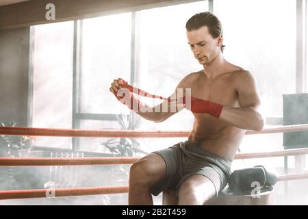 Der junge, gutaussehende Boxer zieht vor dem Kampf oder Training auf dem roten Fass im Boxring. Boxkämpfer Bereiten Verbände Vor Stockfoto