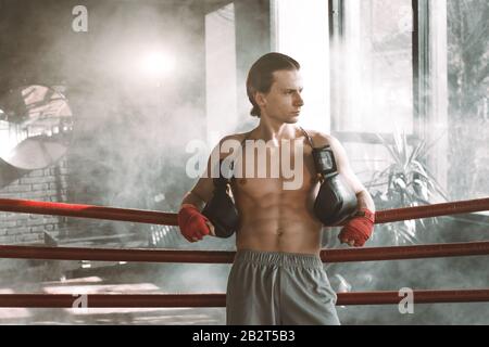 Boxer mit muskulösem Körper, der in einem Boxring steht. Boxer ruht seine Arme auf dem Seil des Boxrings. Stockfoto