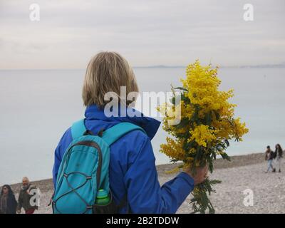 Eine Frau blickt auf das Meer und hält einen großen Haufen gelber Mimosen, die nach der Annullierung der Parade in Nizza verteilt wurden. Stockfoto